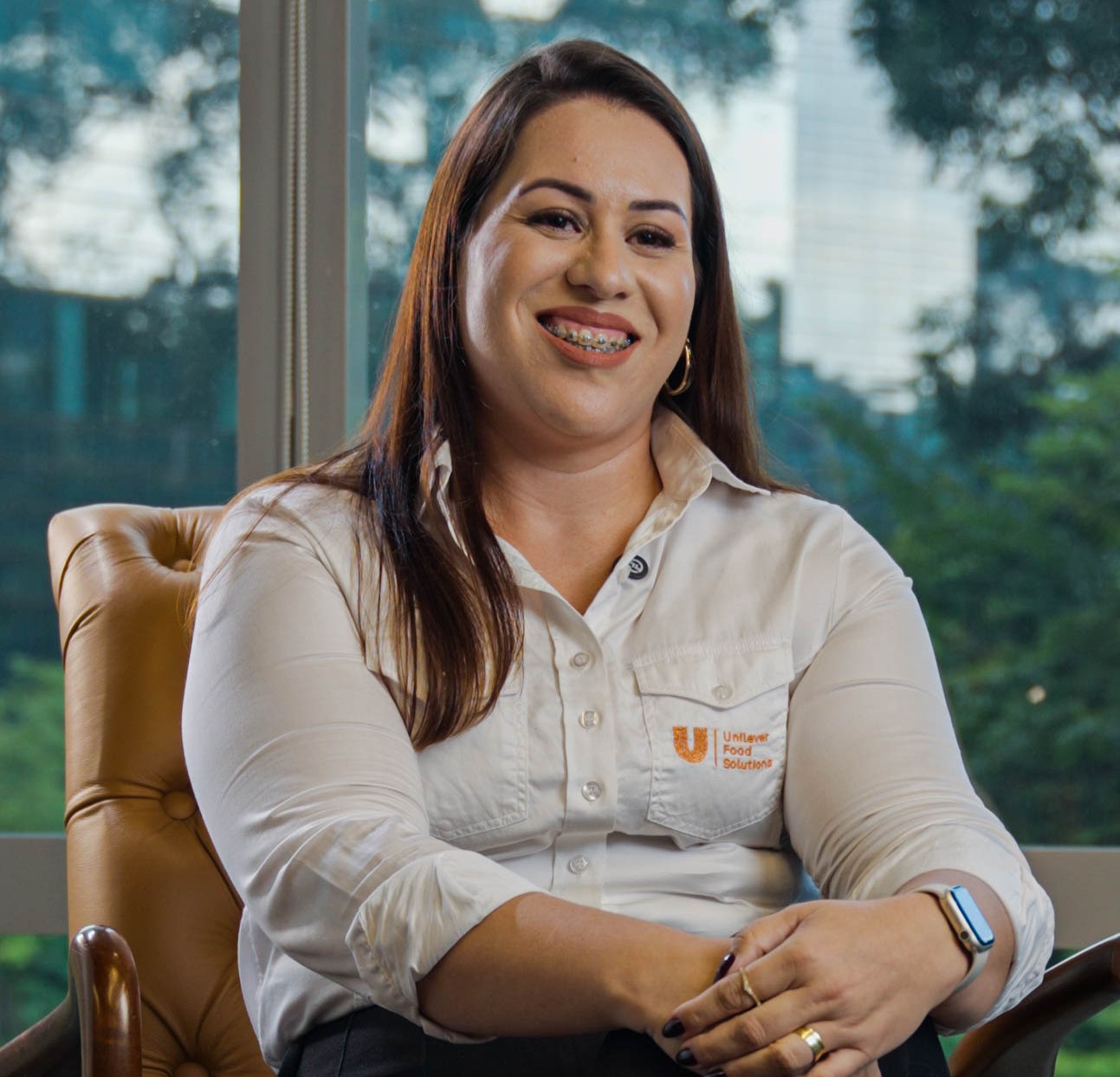 Mulher sorridente sentada em uma poltrona em ambiente corporativo, usando camisa branca com logo da Unilever, com janelas ao fundo e vista para área arborizada