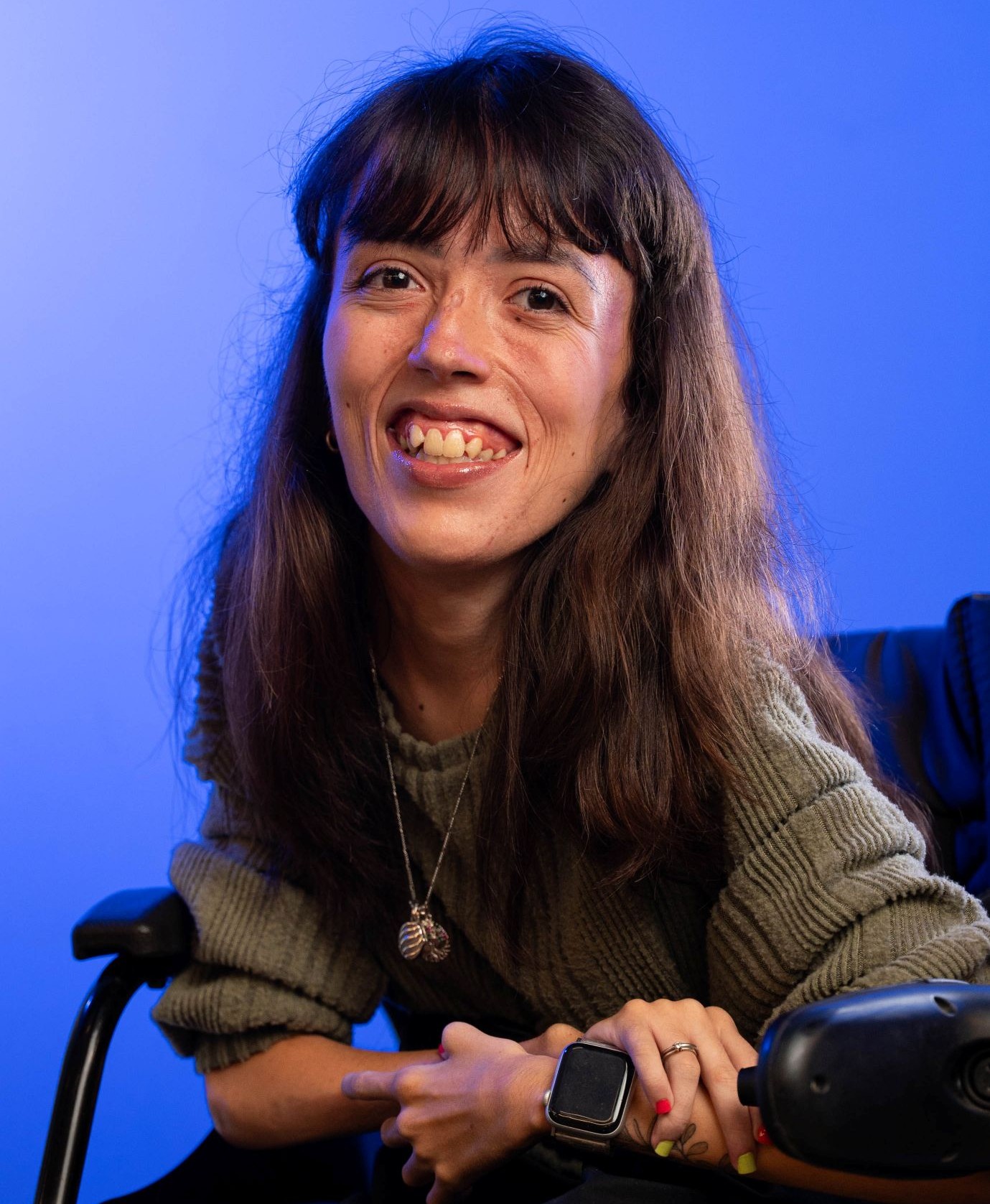 Mulher branca de cabelos castanhos longos que está sorrindo e posa para a foto. Ela usa uma blusa verde e está sentada em uma cadeira de rodas. O fundo é azul.