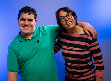 Imagem com fundo azul em que um homem vestindo camisa verde abraça uma mulher de óculos e blusa laranja e preta listrada. Os dois estão sorrindo.
