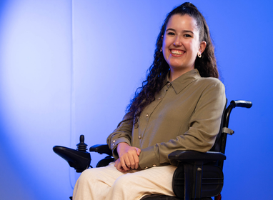 Mulher sorridente sentada em cadeira de rodas motorizada, usando camisa marrom e calça clara, posicionada diante de um fundo azul em degradê.