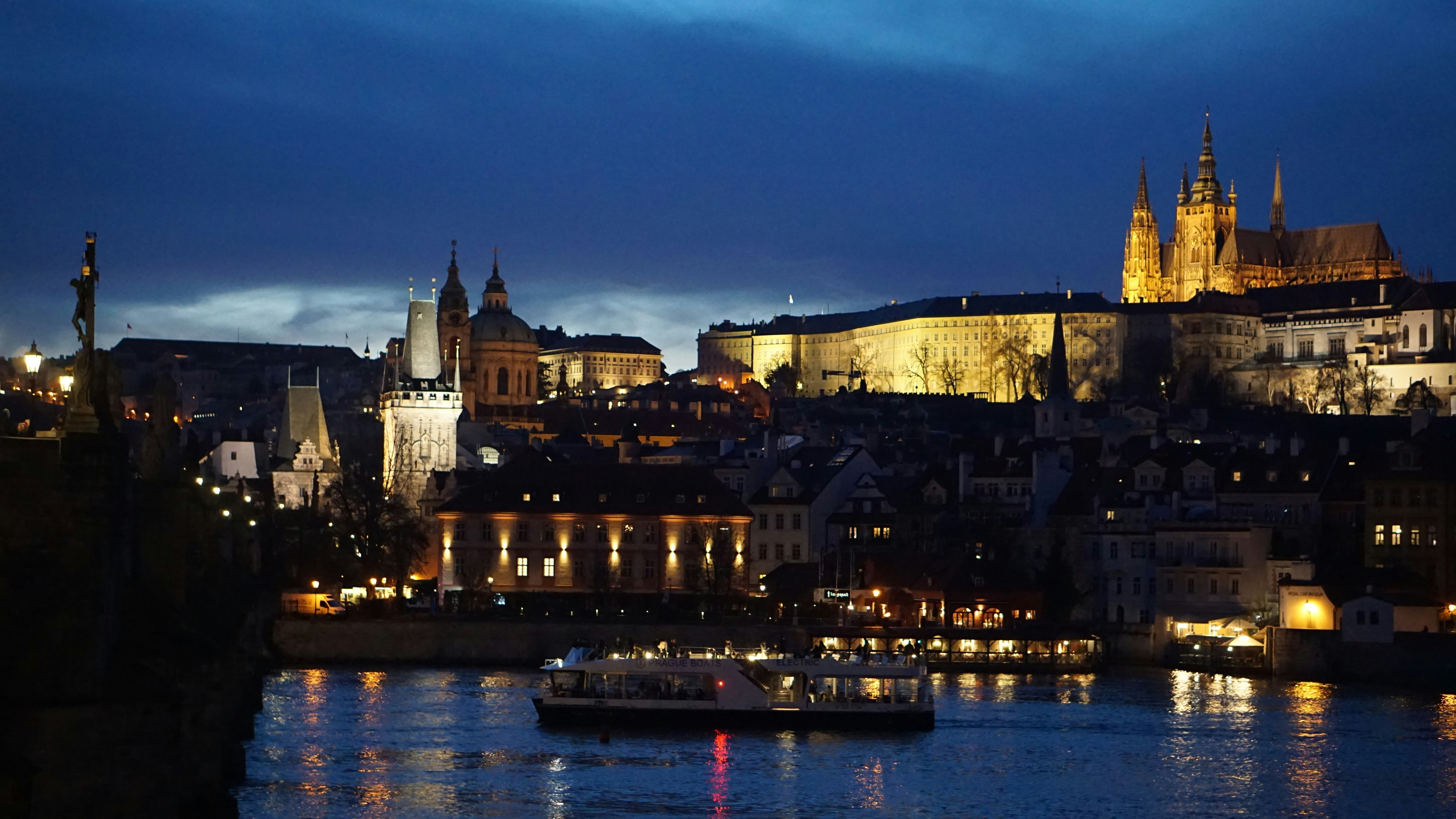 Night view of a riverside cityscape with illuminated historic buildings and a boat on the water.