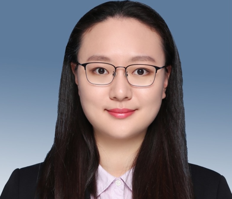 A woman with long dark hair and glasses, wearing a black blazer and light pink collared shirt, pictured against a blue background.