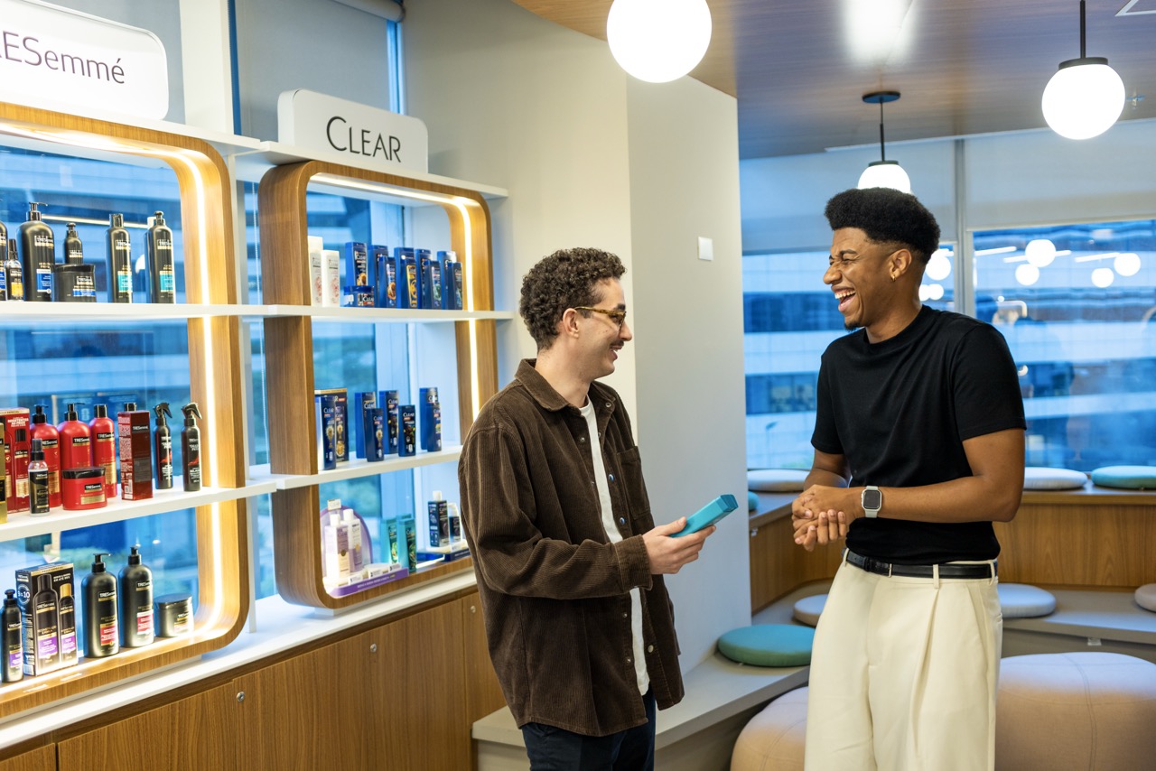 Two Unilever colleagues chat and laugh beside shelves of hair-care products from brands including TRESemmé and CLEAR.