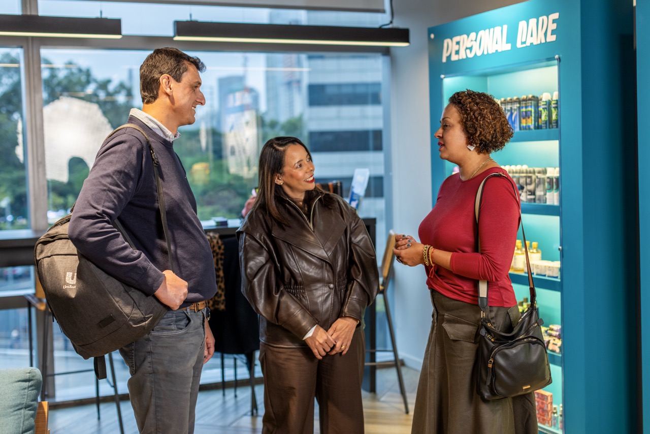 Three colleagues smile and chat together in a bright Unilever office, surrounded by displays of personal care and beauty products.