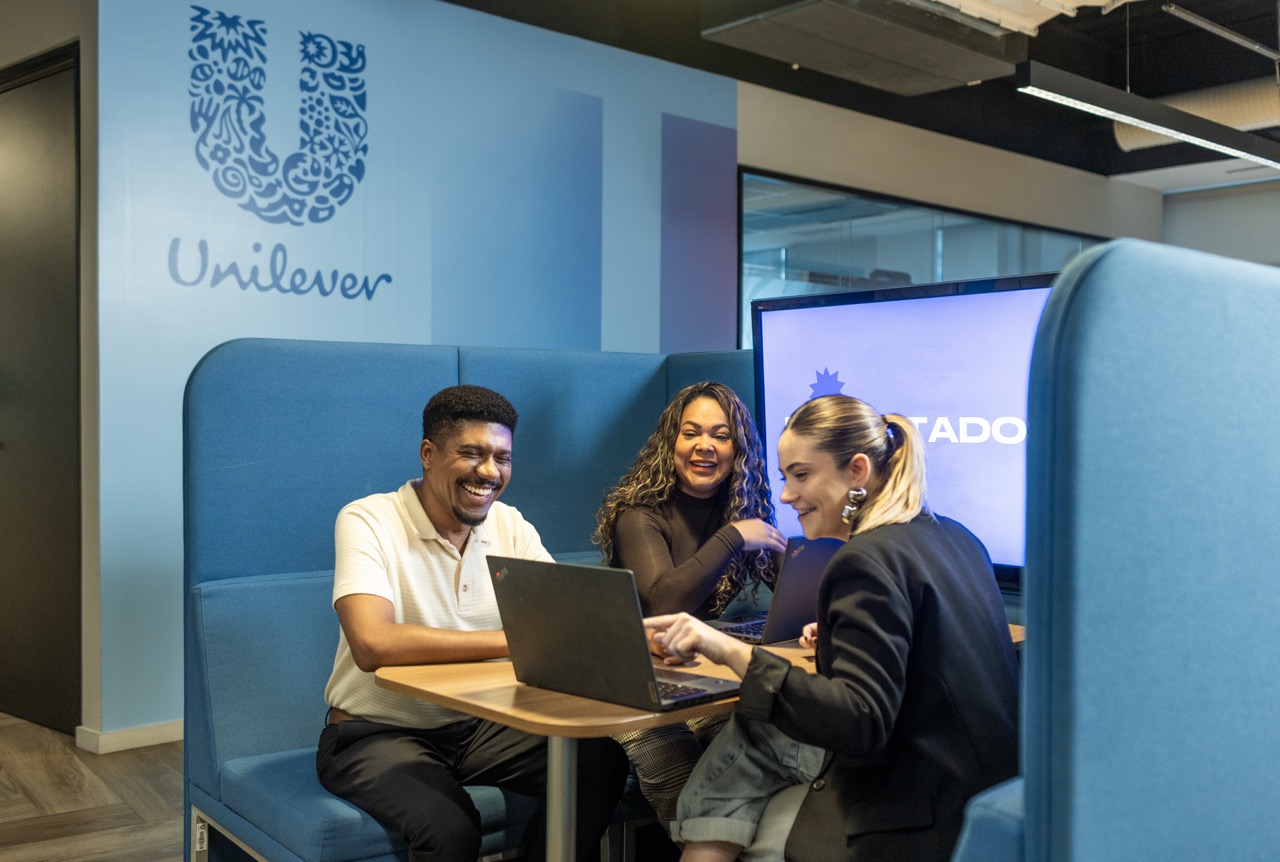 Three Unilever colleagues smile and collaborate around laptops in a comfortable office seating area.