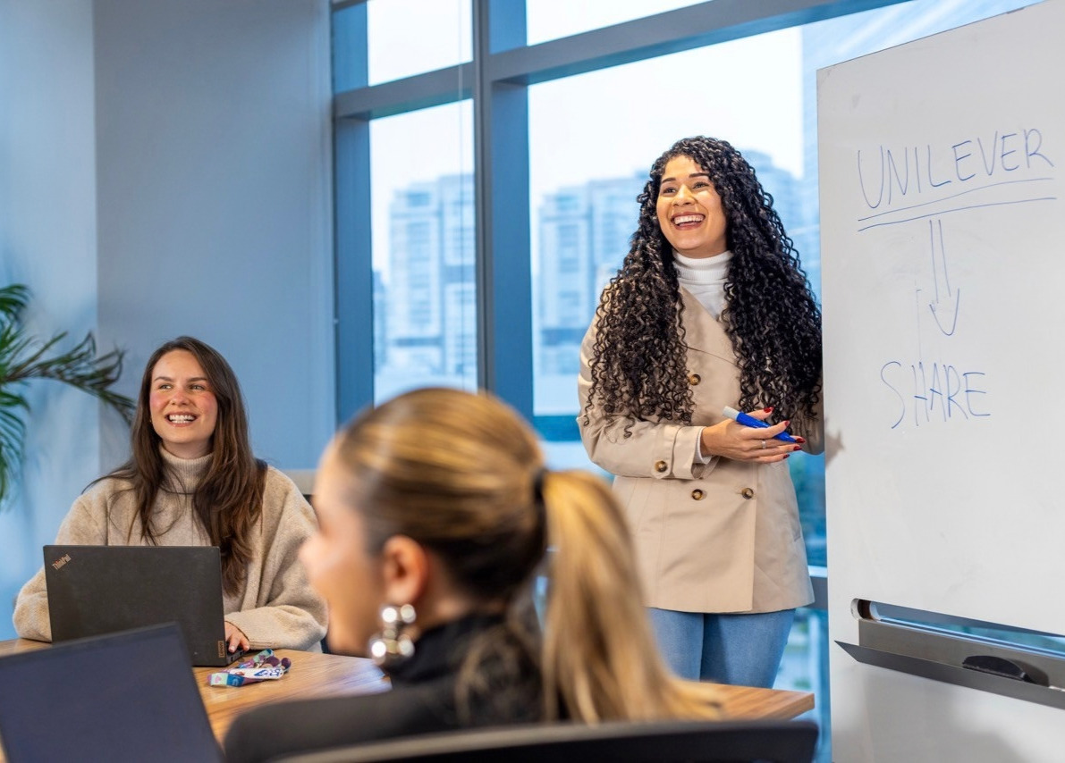 Three colleagues in a bright meeting room smile as one presents at a whiteboard showing "UNILEVER" with an arrow pointing down to "SHARE".