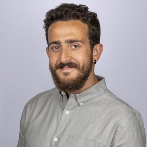 smiling man with curly dark hair and a beard, wearing a light grey button-down shirt, pictured against a pale grey background.