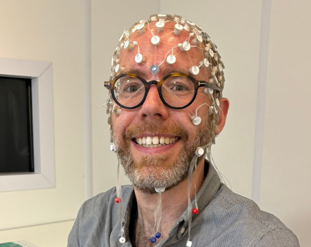 A smiling man wearing glasses and a cap fitted with numerous electrodes, with additional electrodes attached to his face and neck.