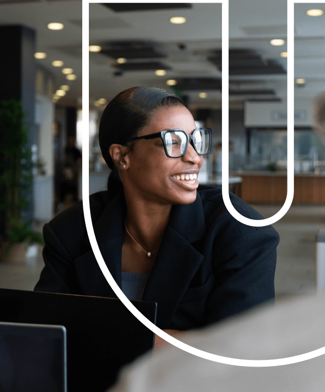 Female professional in an open office settings looking out of the window smiling. There is an overlay of a white letter in the shape o of a capital U