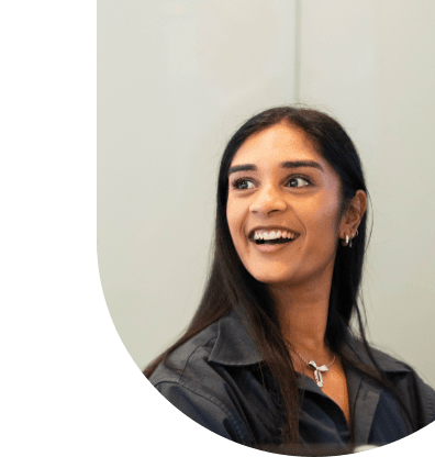 Smiling woman with long dark hair wearing a cream-colored top, standing indoors in a bright office environment.