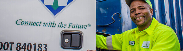 Smiling male employee, wearing yellow shirt and cap, opening a door with Waste Connections logo and tagline on it.
