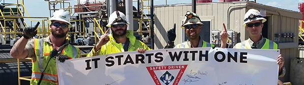 Four male employees wearing hard hats, sunglasses and yellow safety vests, holding a sign that says: 'It starts with one.'