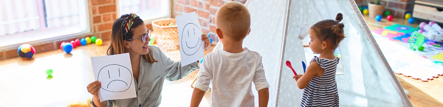 Erzieherin spielt mit zwei kleinen Kindern und zeigt einen weinenden und einen lächelnden Smiley auf  einem weißen Blatt Papier. 
