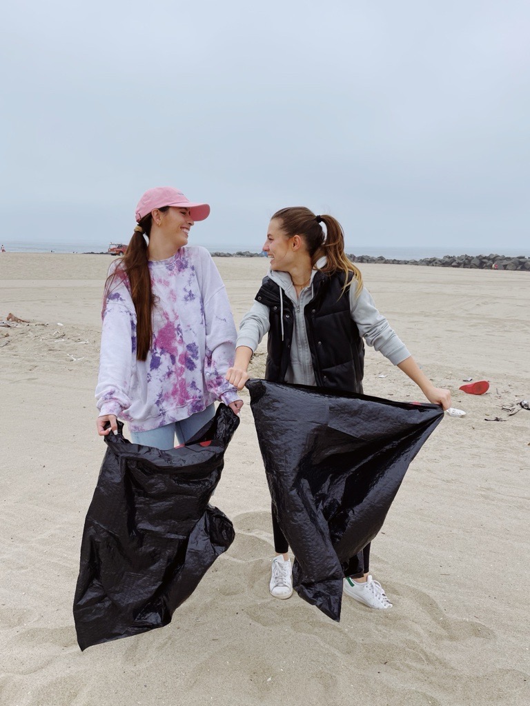 Athena and another woman stand on a beach holding trash bags.