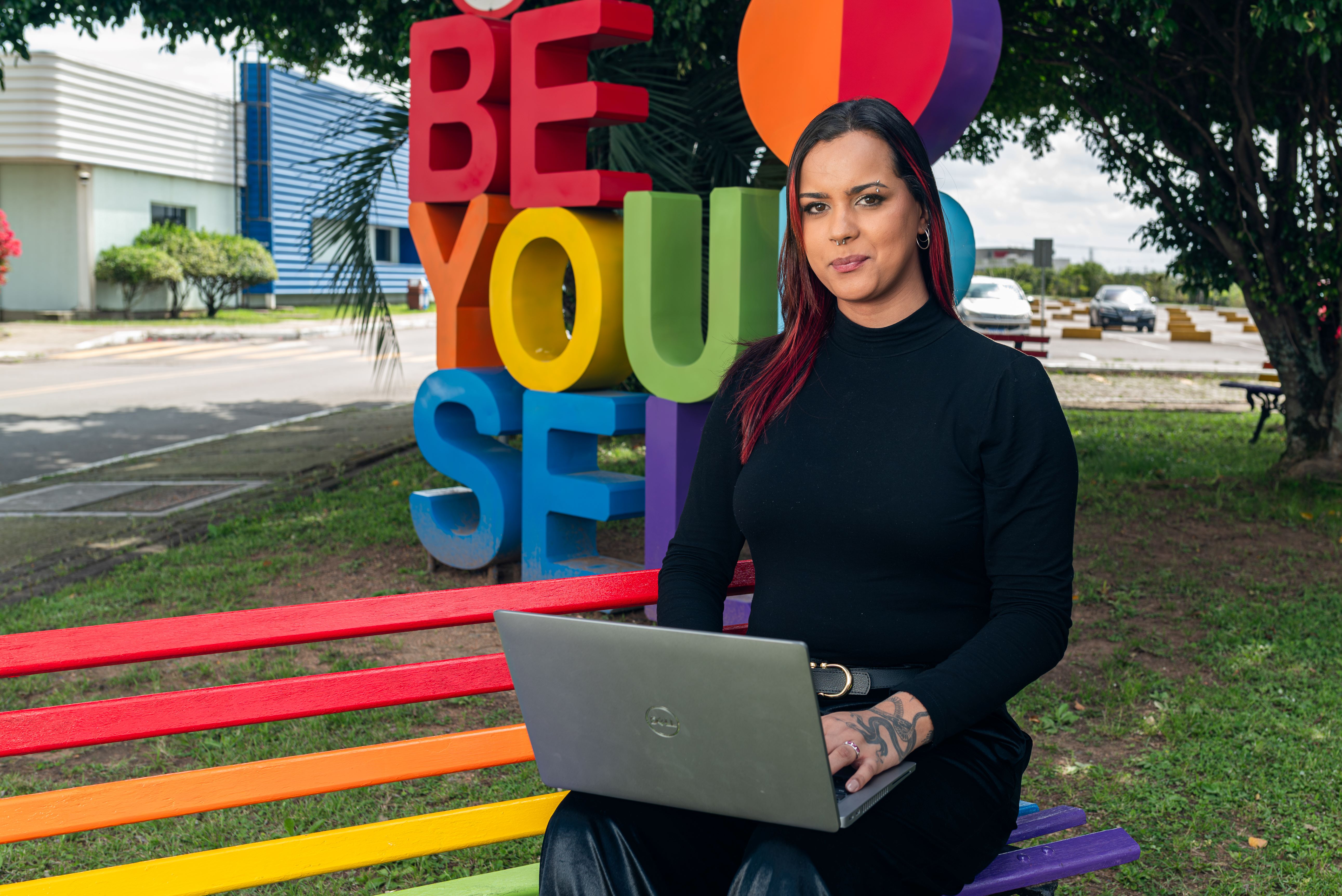 Agni sits on a rainbow bench with a sigh behind her that reads, "Be your self" in rainbow letters.
