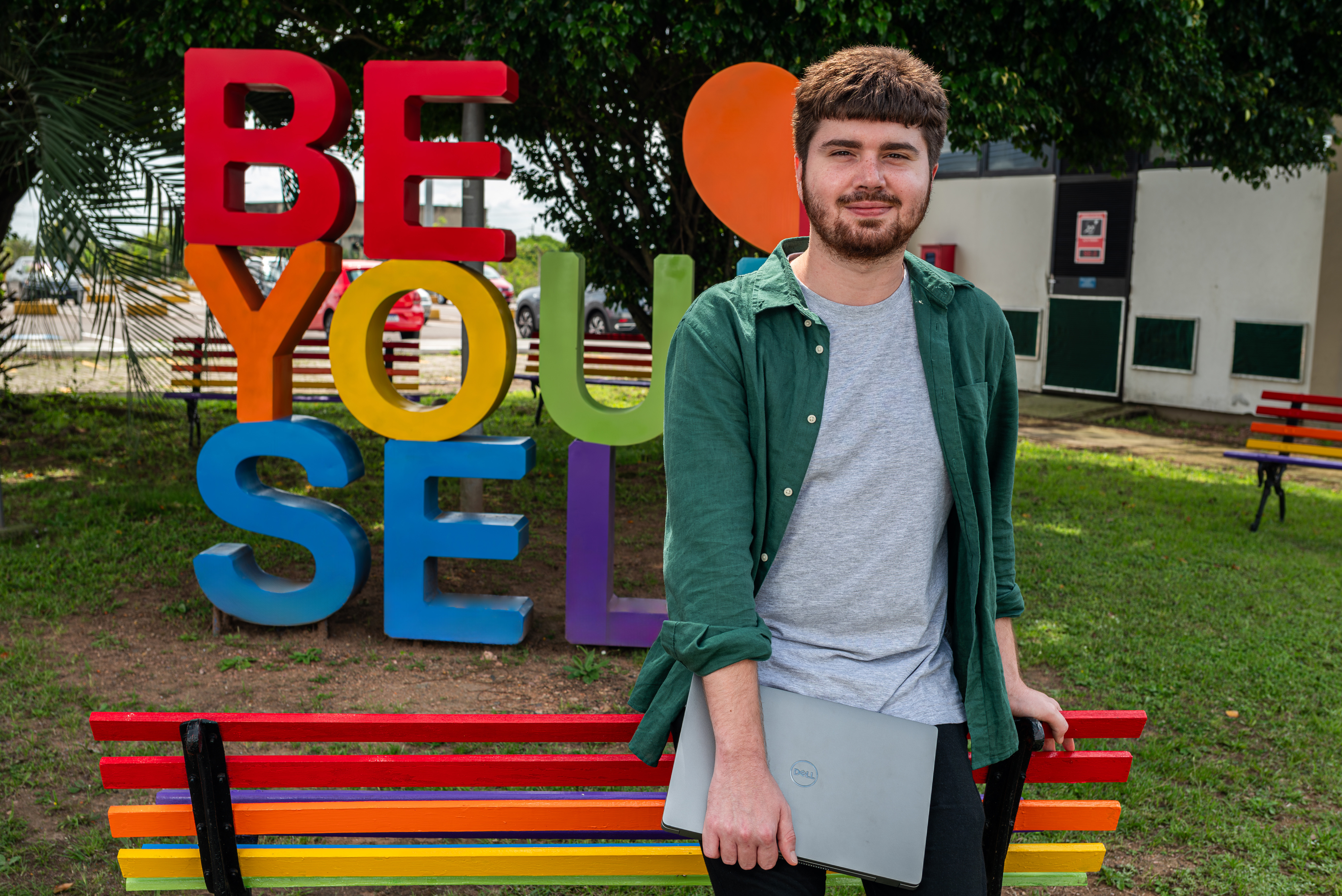 Guilherme smiles in front of a rainbow sign that says, "Be Your Self."