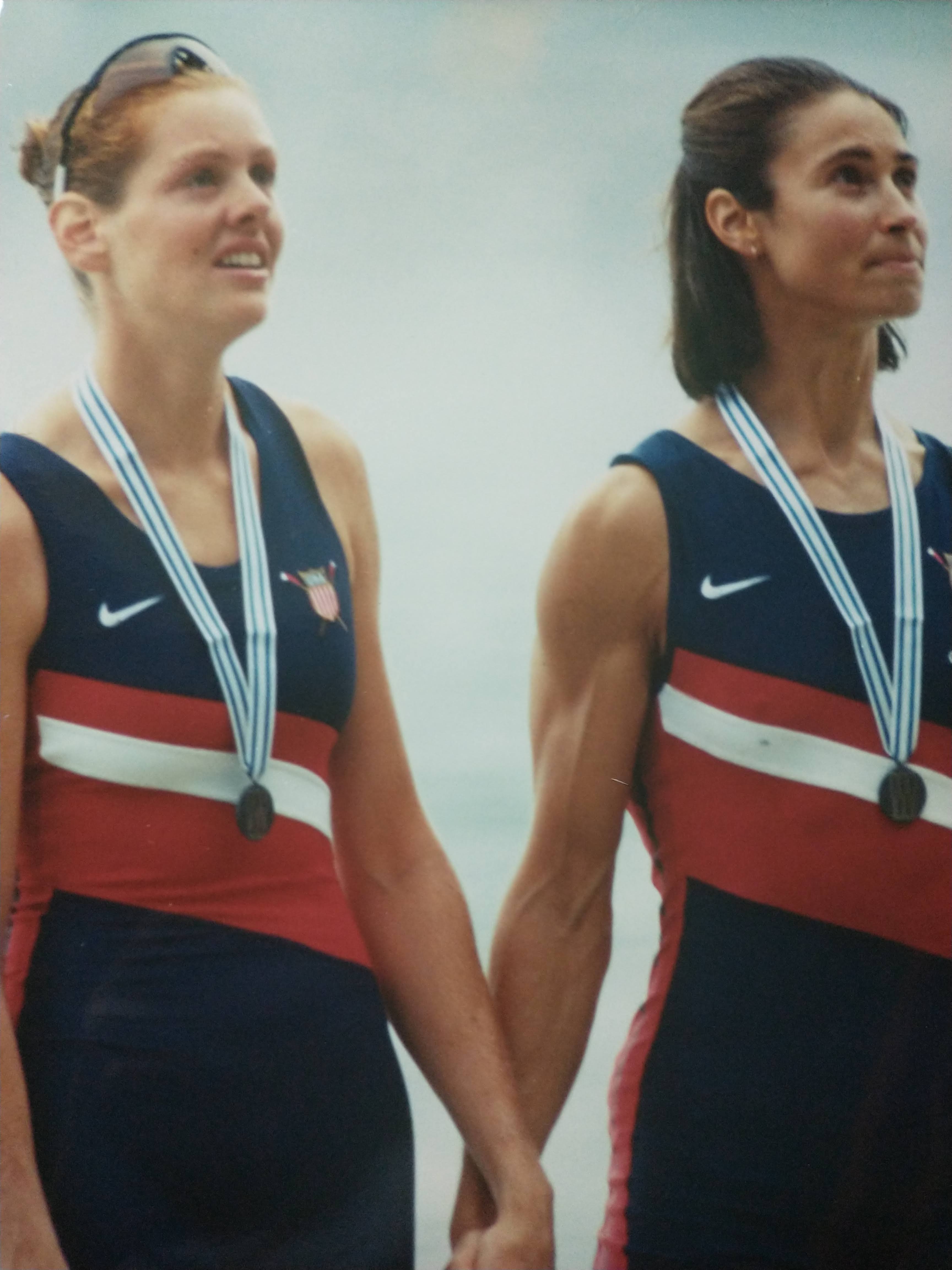 Michelle, dressed in United States rowing gear, wears a medal and holds hands with a rowing teammate.