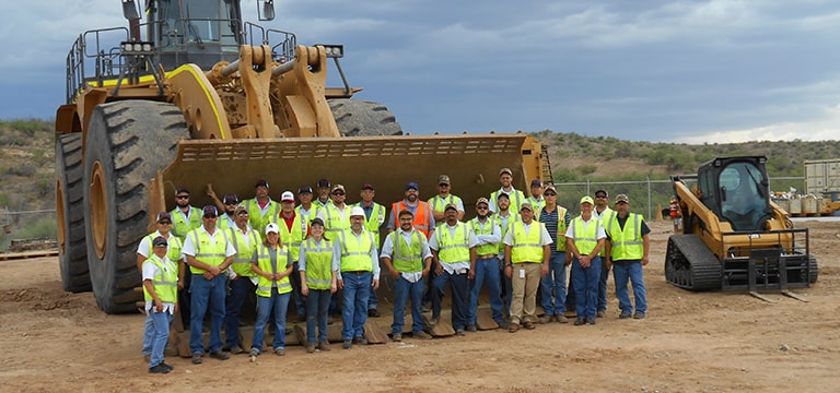 Gran grupo de hombres y mujeres de pie frente a una gran excavadora y topadora mecánica