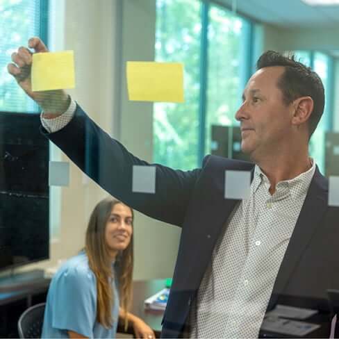 Male employee putting sticky notes on a glass wall while another employee looks on