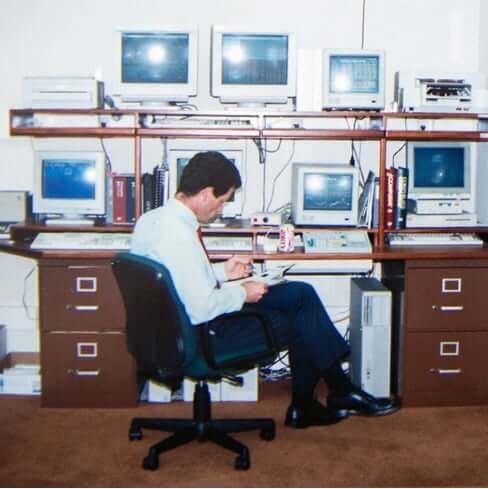 Throwback image of Ken Fisher sitting at his desk looking at notes, surrounded by several computers