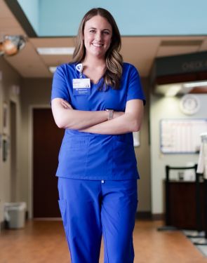 Female nurse in blue scrubs stands in a hospital hallway with arms crossed.