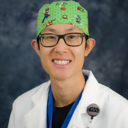 Andrew smiles in a scrub hat and white coat with glasses.