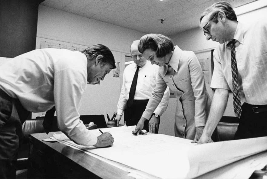 Black and white photo of Katharine Graham and three male colleagues in an office standing over a large piece of paper.