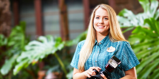 Woman, with long blonde hair, wearing a blue uniform, smiling and holding a camera.