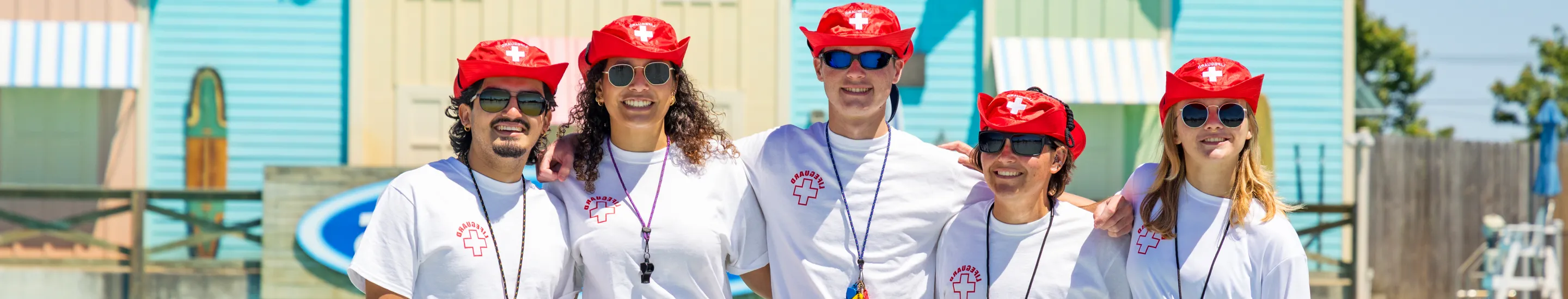 Five smiling lifeguards wearing white t-shirts and red hats.