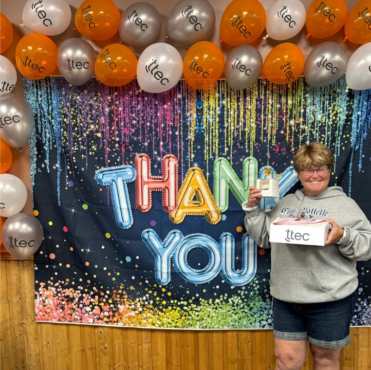 Happy TTEC team member celebrating in front of colorful "THANK YOU" backdrop and TTEC balloons.