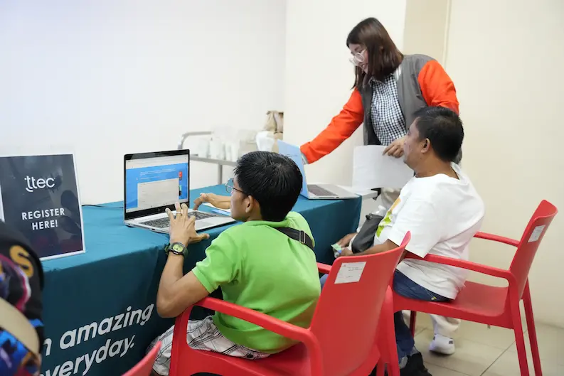 A recruiter assists persons with disabilities at a job fair booth, guiding them through the application process using printed materials and a laptop.