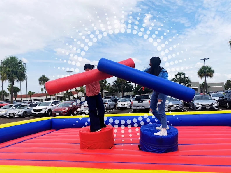 Two people balancing on an inflatable platform while playfully battling each other with large foam logs