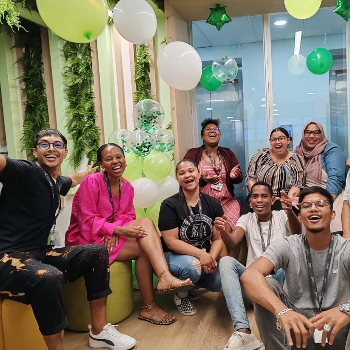 Group of cheerful employees sitting and posing together in a brightly decorated office space with green and white balloons, leafy wall decor, and festive hanging ornaments.