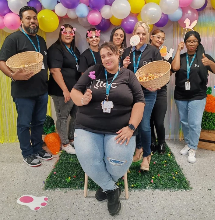 Group of people in matching TTEC shirts posing with baskets and festive props in front of a colorful balloon backdrop.