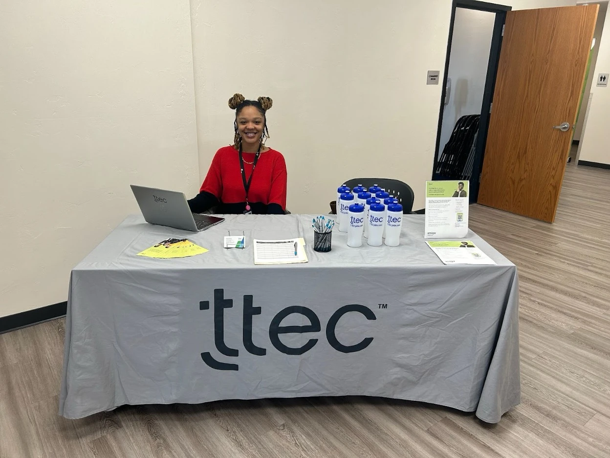 TTEC employee sitting at a branded recruitment table with laptop, promotional materials, and water bottles.