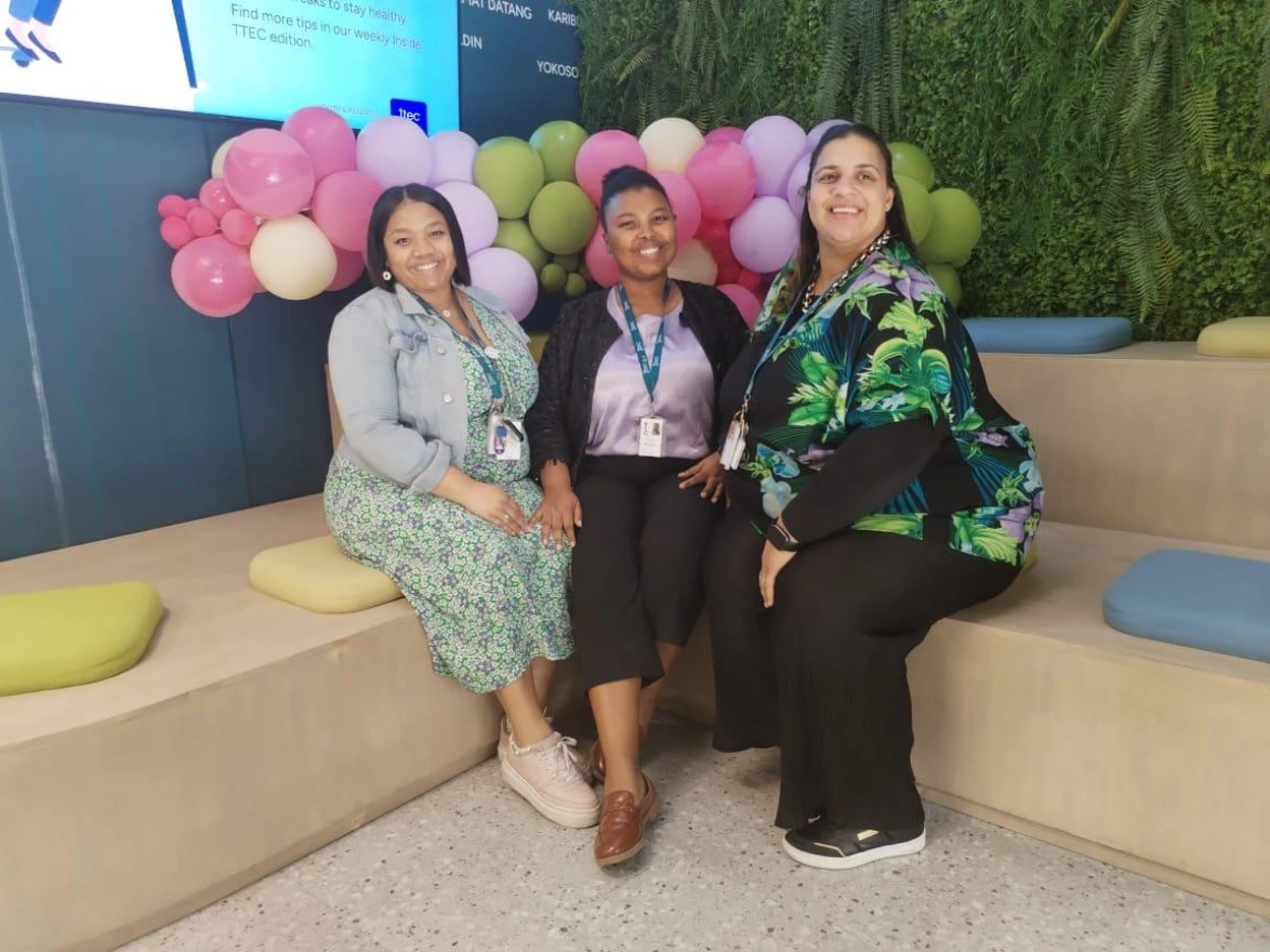 Three TTEC employees sitting together with colorful balloons in the background.