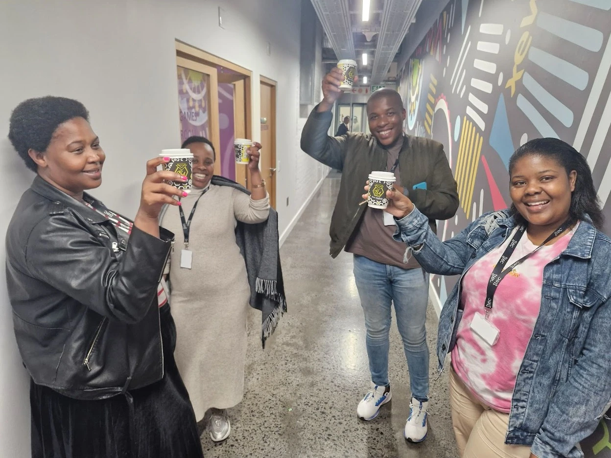 Four TTEC employees smiling and raising coffee cups in a celebratory toast in an office hallway.