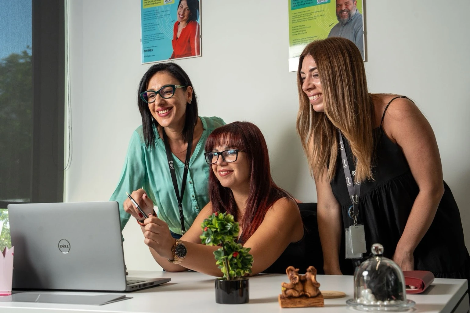 Three female colleagues smiling while gathered around a laptop computer in a modern office setting.