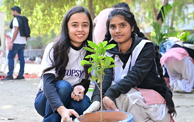 Two young girls kneeling down outside and smiling while posing with a plant.