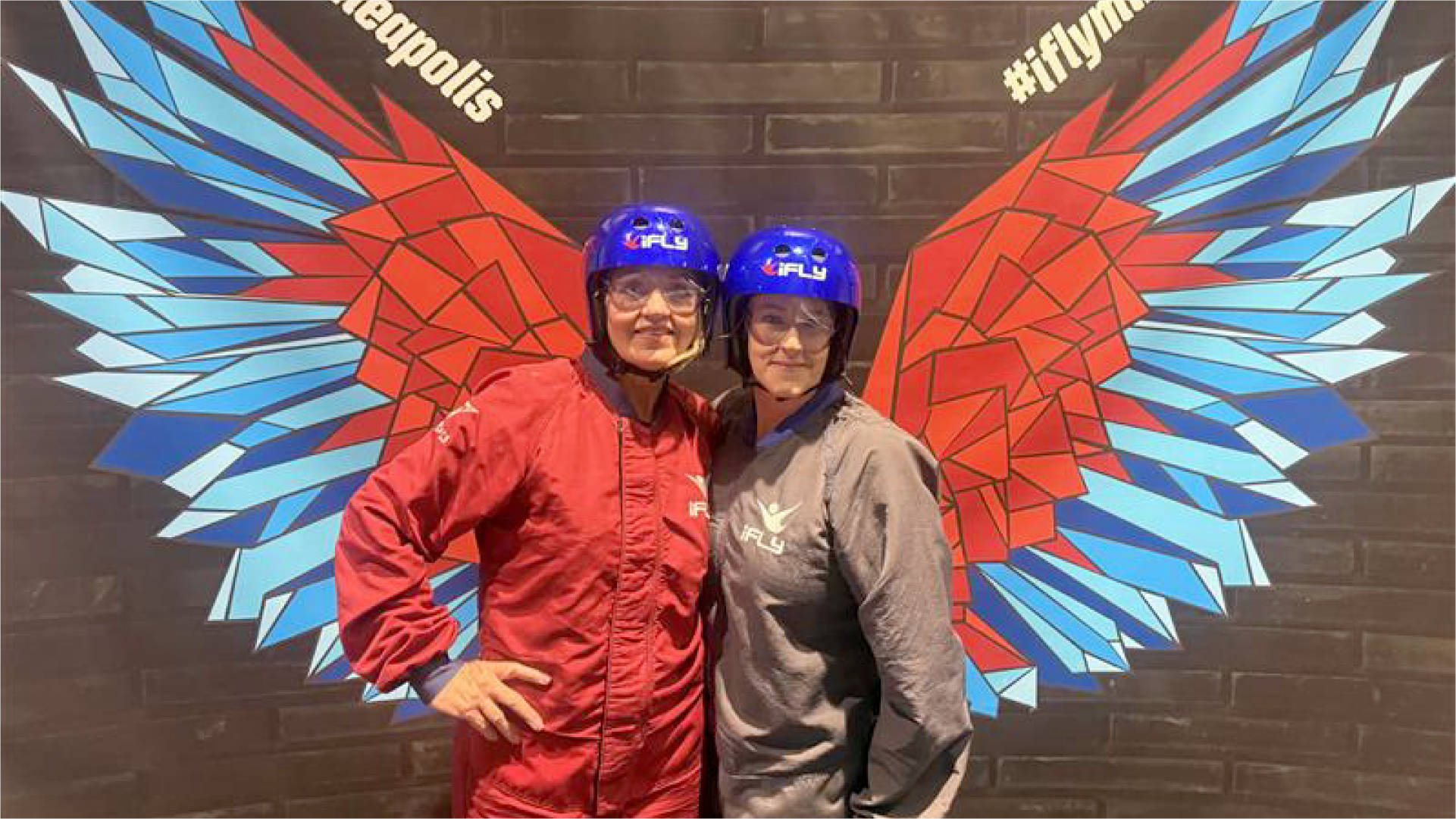 Tami and her franchisee Kim stand in front of a winged selfie backdrop at iFly, a skydiving simulation experience