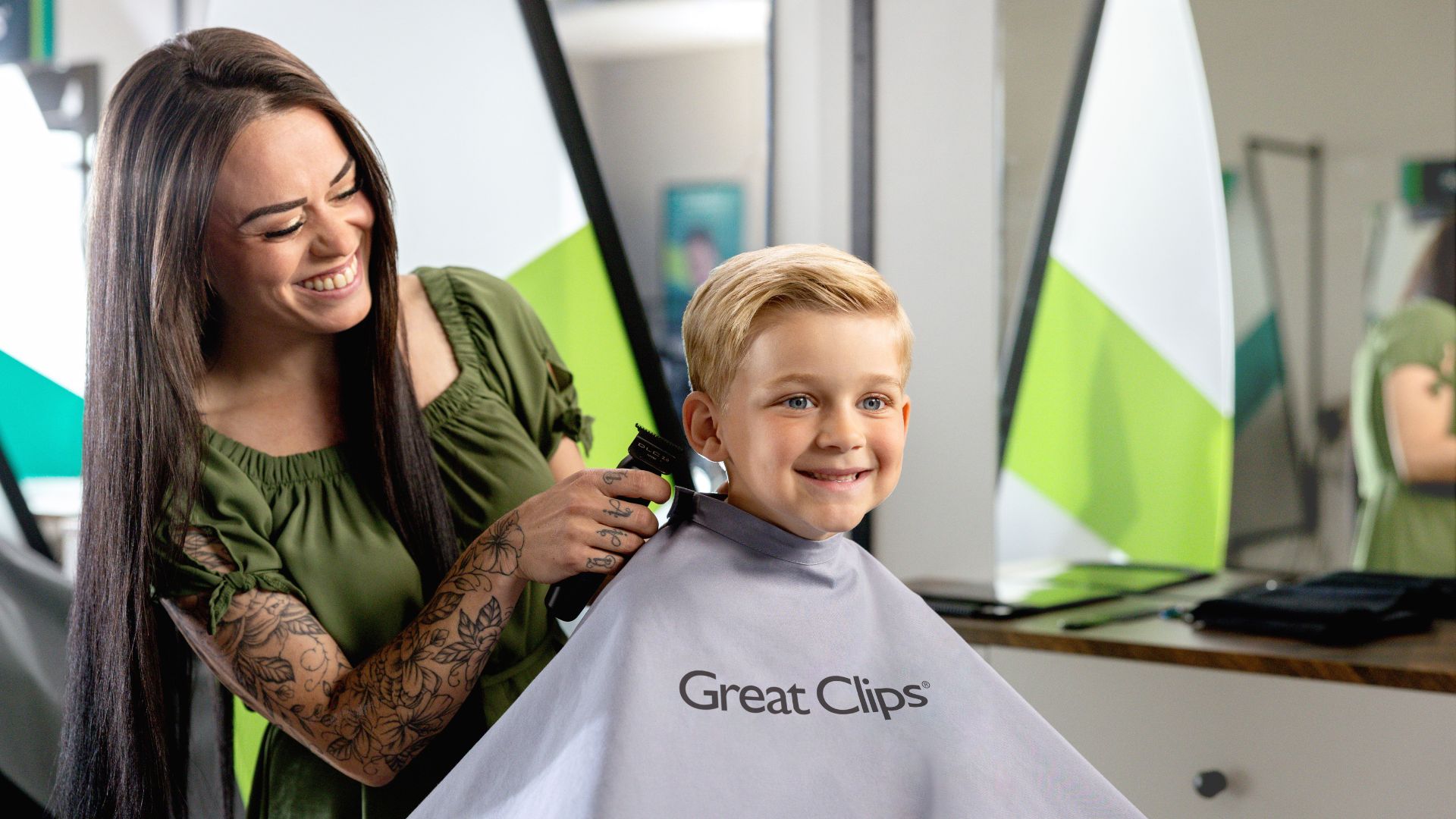 Smiling stylist with long, straight brown hair and tattoos cuts the hair of young smiling boy with blond hair.