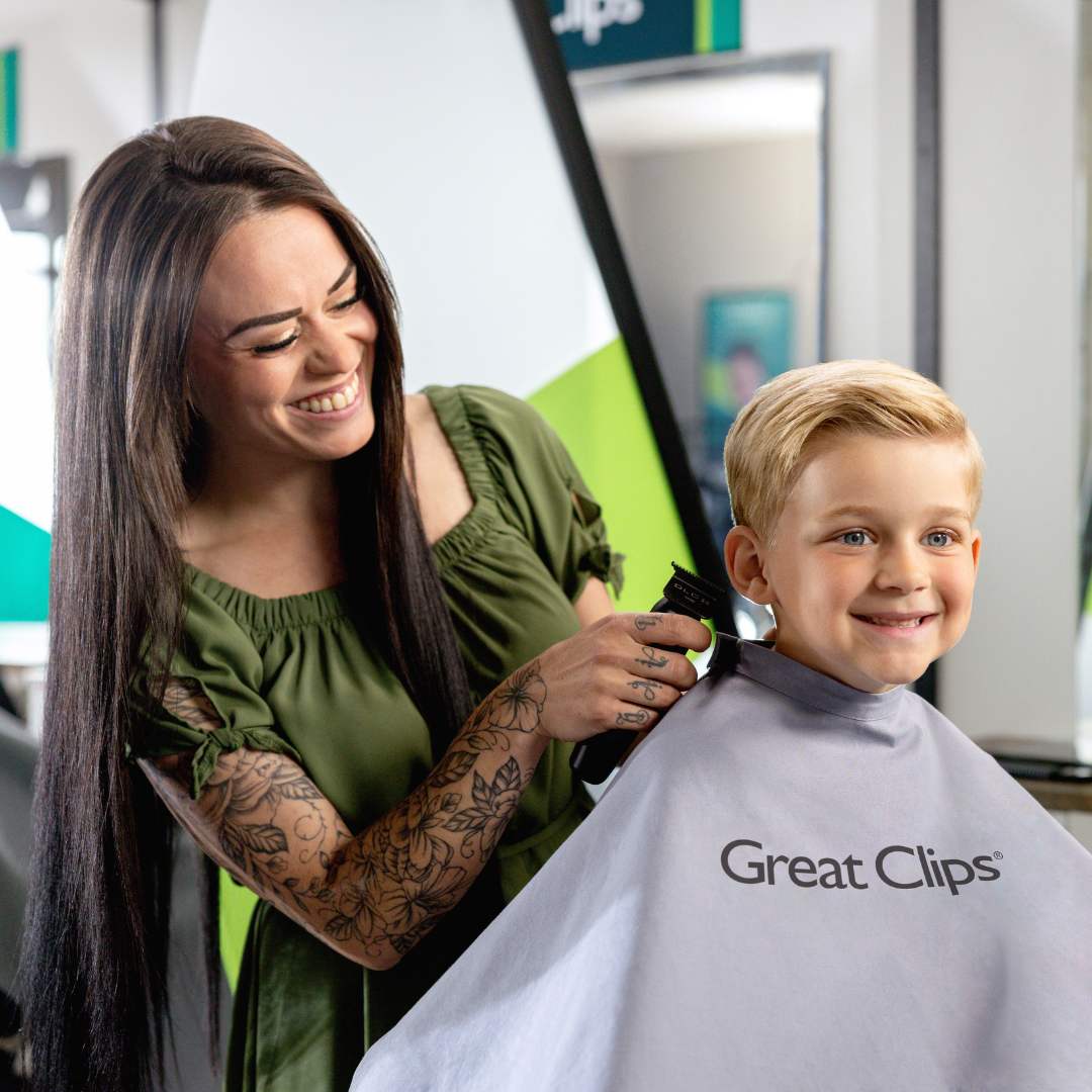 Smiling stylist with long, straight brown hair and tattoos cuts the hair of young smiling boy with blond hair.