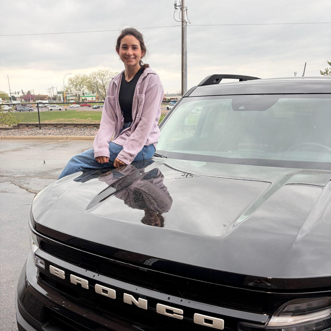 Natalia sits on the hood of her new Bronco, smiling.