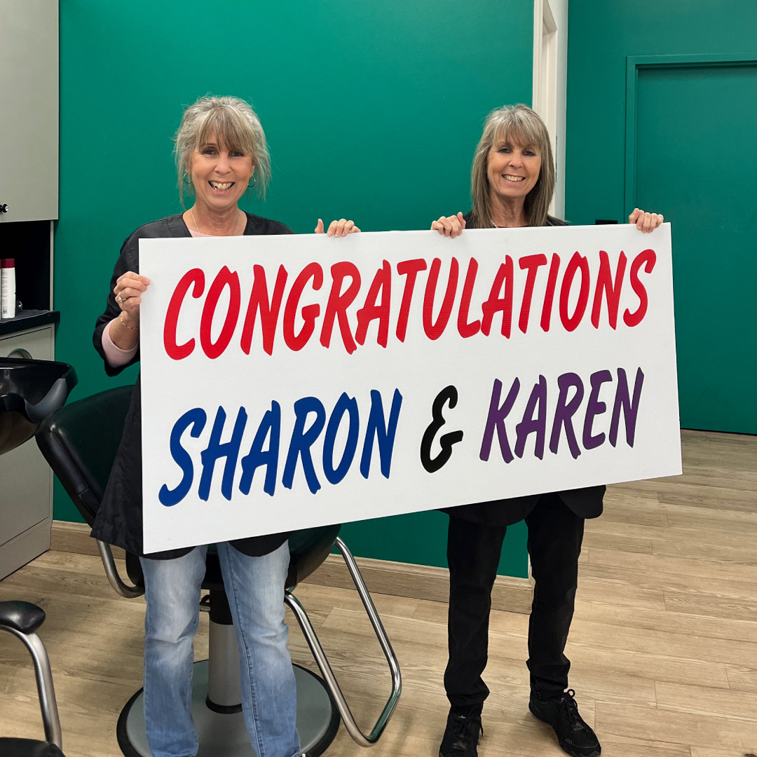 Smiling twin stylists holding a sign that says "Congratulations Sharon and Karen"