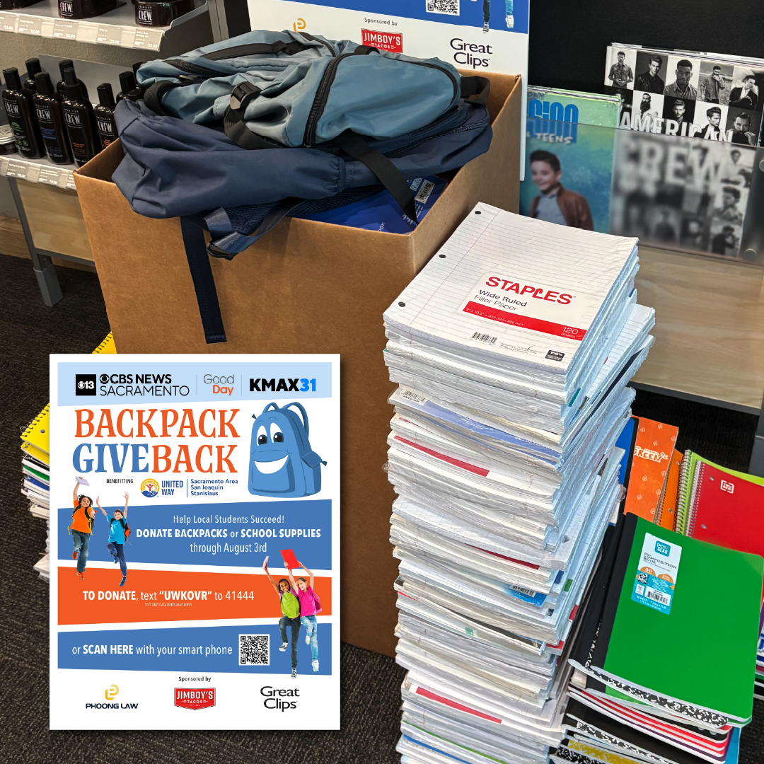 A stack of school supplies sits next to a box full of backpacks in a Great Clips lobby.