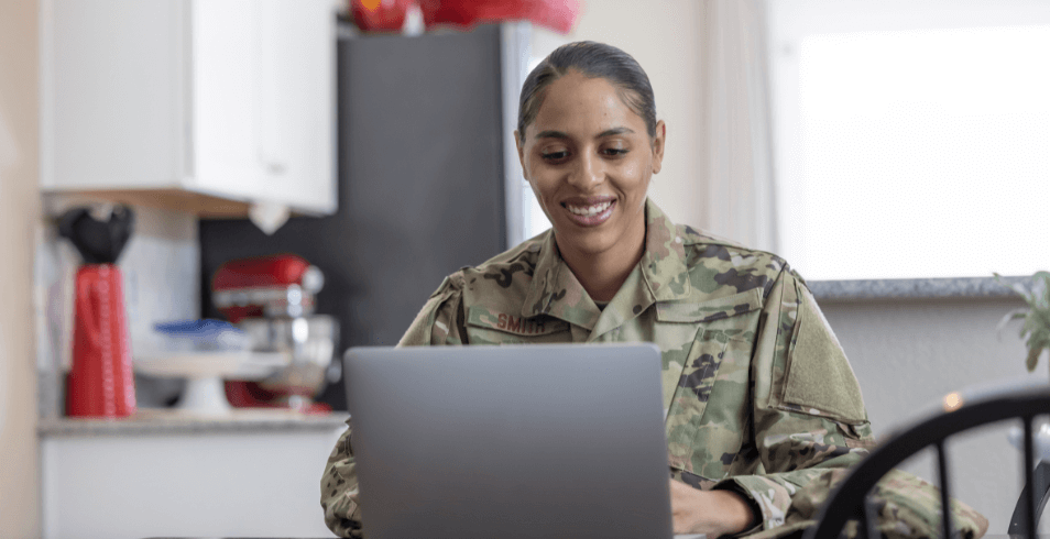 female service member in military fatigues, sitting behind a laptop and smiling