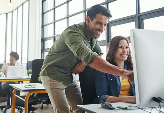 smiling man standing over the shoulder of a woman in a large, sunlit office, pointing at her desktop computer screen