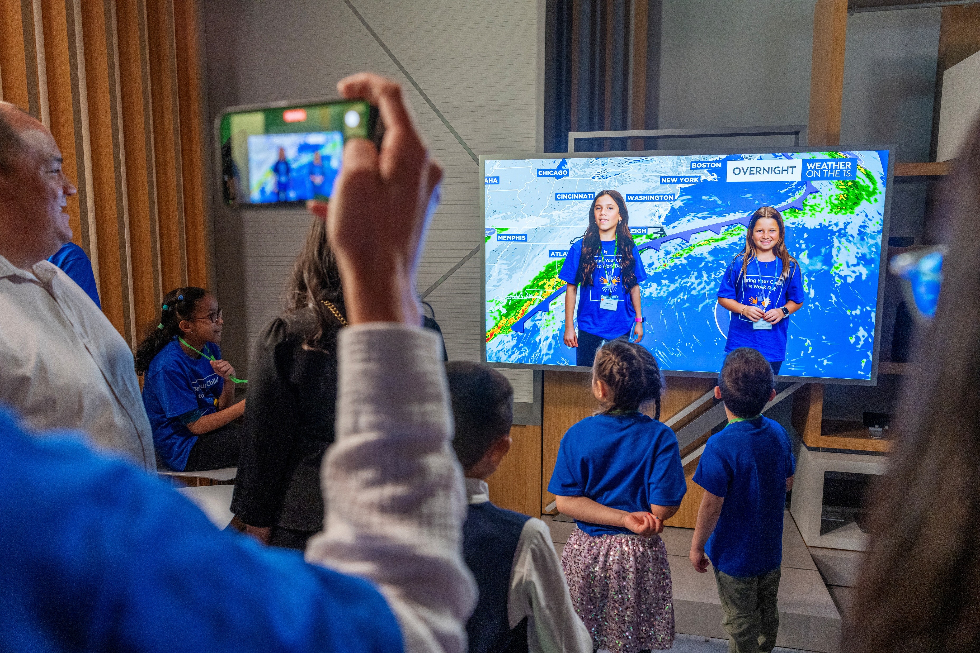 children gather to watch monitor featuring 2 young girls conducting a faux weather forecast