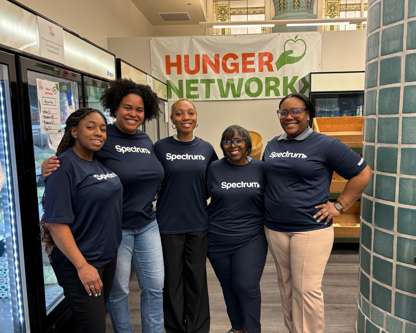 5 women in Spectrum blue t-shirts pose next to freezer near Hunger Network sign
