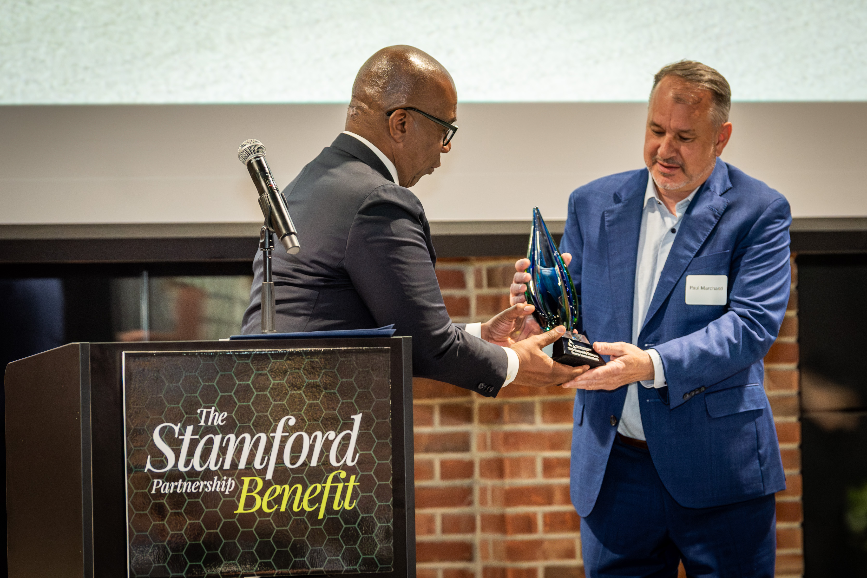 Darrell Johnson, an African American man in a gray suit, presents award to Paul Marchand, a white man in a blue suit